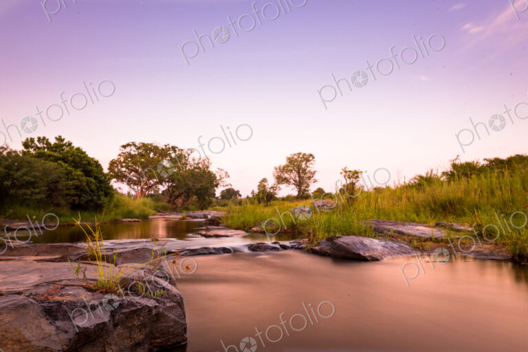 A river flowing over rocks, surrounded by boulders and green grass, pink sky…