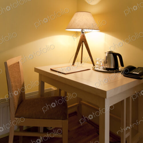 A room with a small desk and chair with a lit table lamp, tray and kettle and telephone.