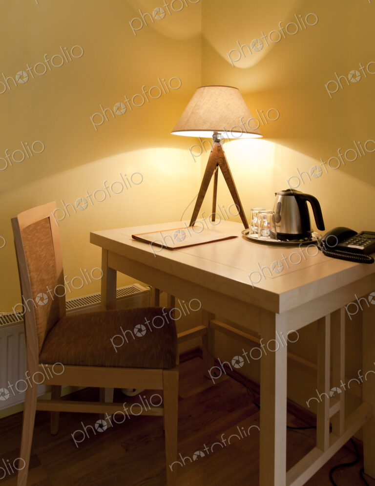 A room with a small desk and chair with a lit table lamp, tray and kettle and telephone.
