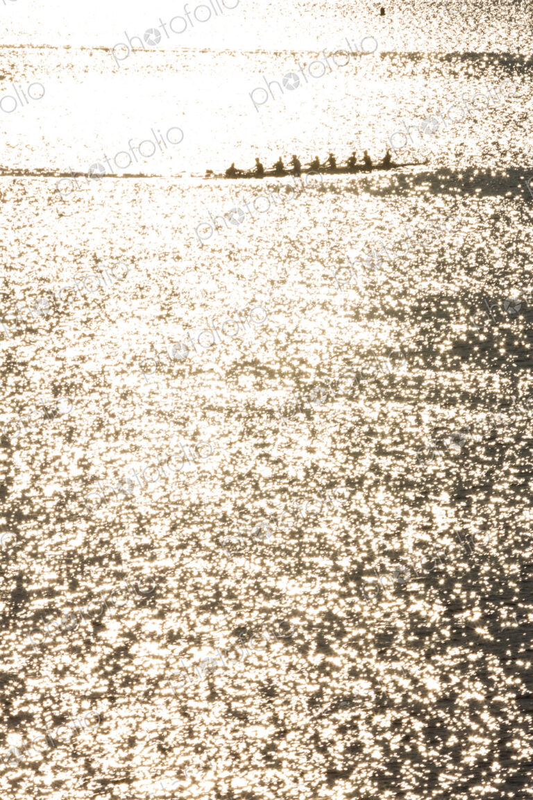 A rowing eight crew in a shell on the water, sunshine reflecting on the surface.