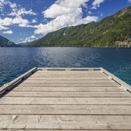 A rustic wooden dock extends over Lake Crescent and its vivid blue water, framed by…
