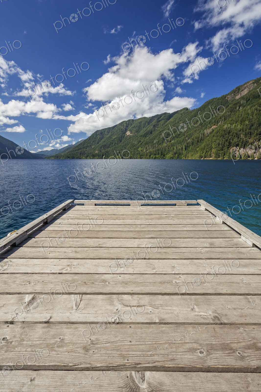 A rustic wooden dock extends over Lake Crescent and its vivid blue water, framed by…