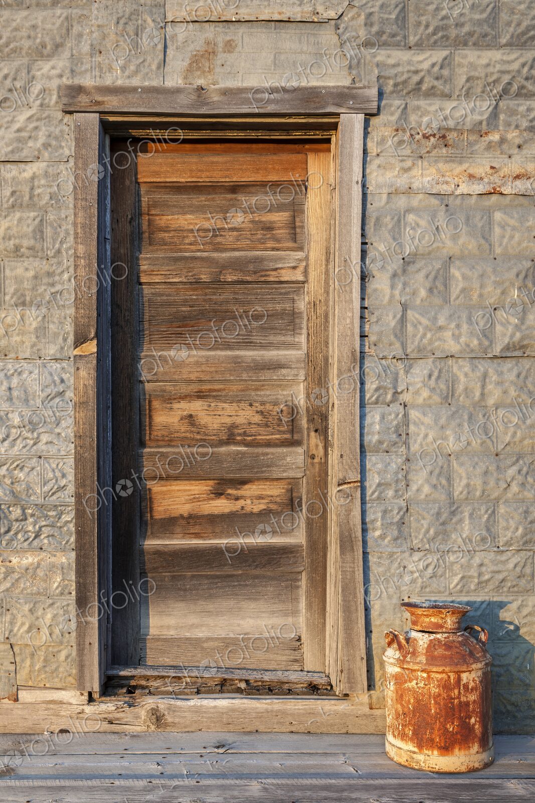 A rustic wooden door and old milk can catch warm evening light on a prairie building…