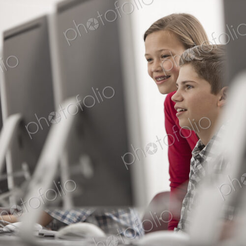 A school computer lab room, with rows of screens. Two young people looking intently at…