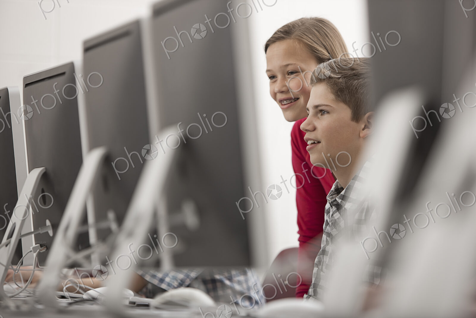 A school computer lab room, with rows of screens. Two young people looking intently at…