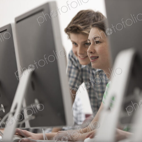 A school computer lab room, with rows of screens. Two young people looking intently at…