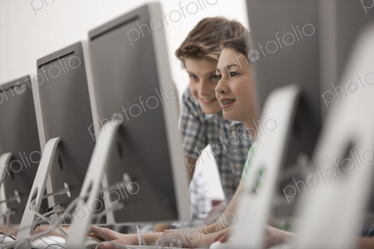 A school computer lab room, with rows of screens. Two young people looking intently at…