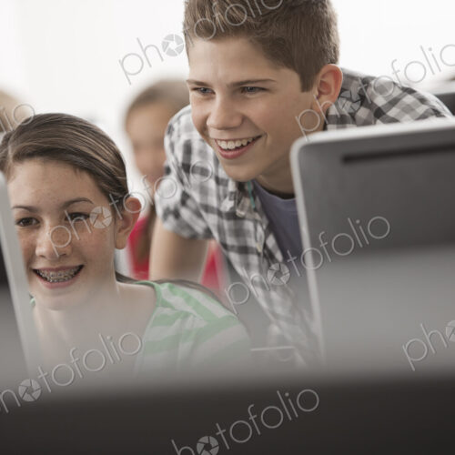 A school computer lab room, with rows of screens. Two young people looking intently at…