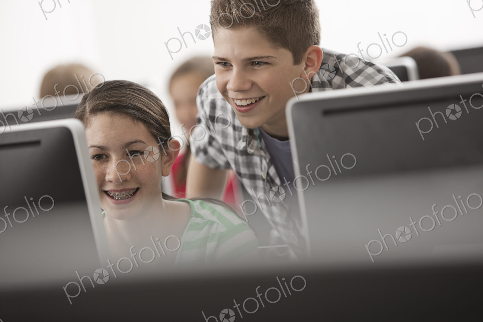 A school computer lab room, with rows of screens. Two young people looking intently at…