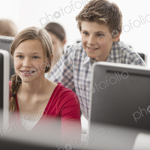 A school computer lab room, with rows of screens. Two young people looking intently at…