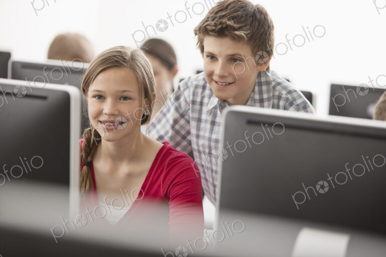 A school computer lab room, with rows of screens. Two young people looking intently at…