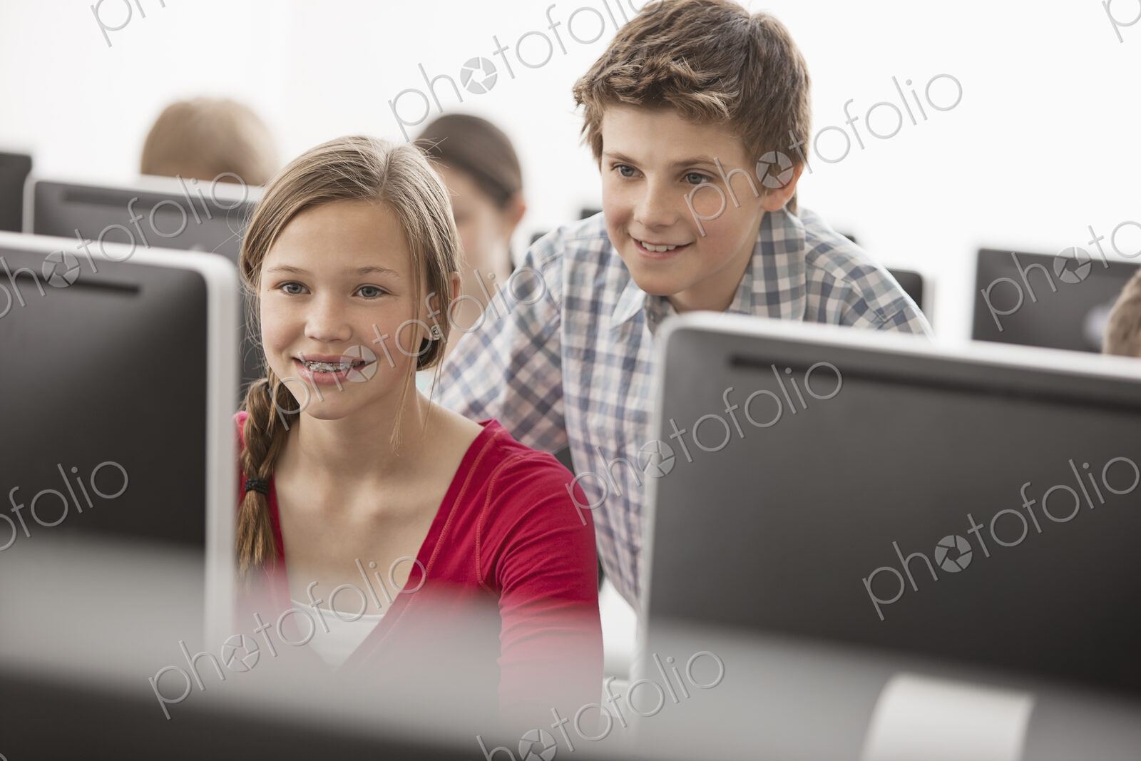 A school computer lab room, with rows of screens. Two young people looking intently at…
