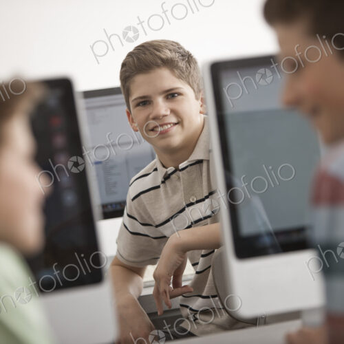 A school room computer lab with rows of computer monitors and seating. Three young…