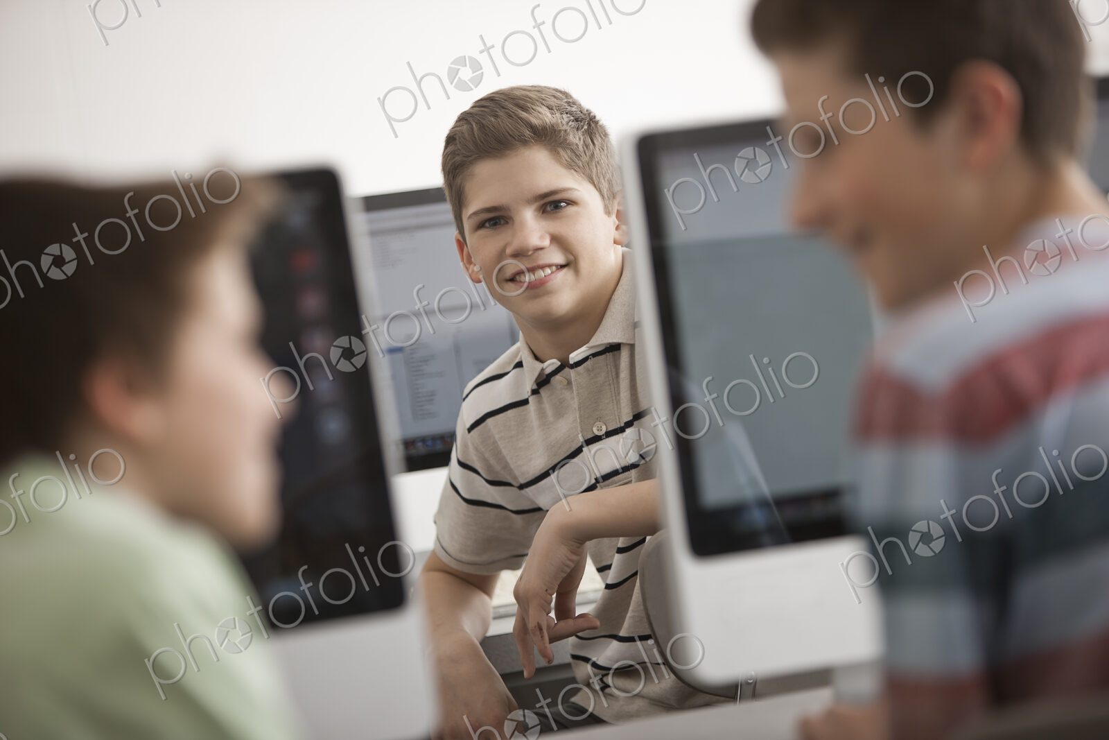 A school room computer lab with rows of computer monitors and seating. Three young…
