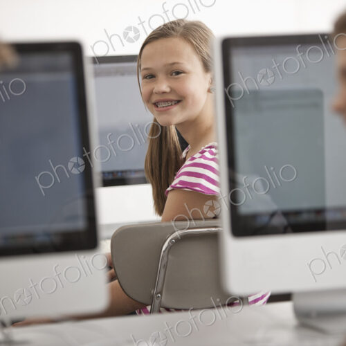A school room computer lab with rows of computer monitors and seating. Three young…