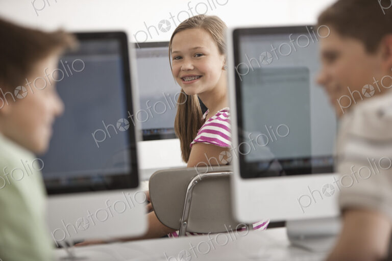 A school room computer lab with rows of computer monitors and seating. Three young…