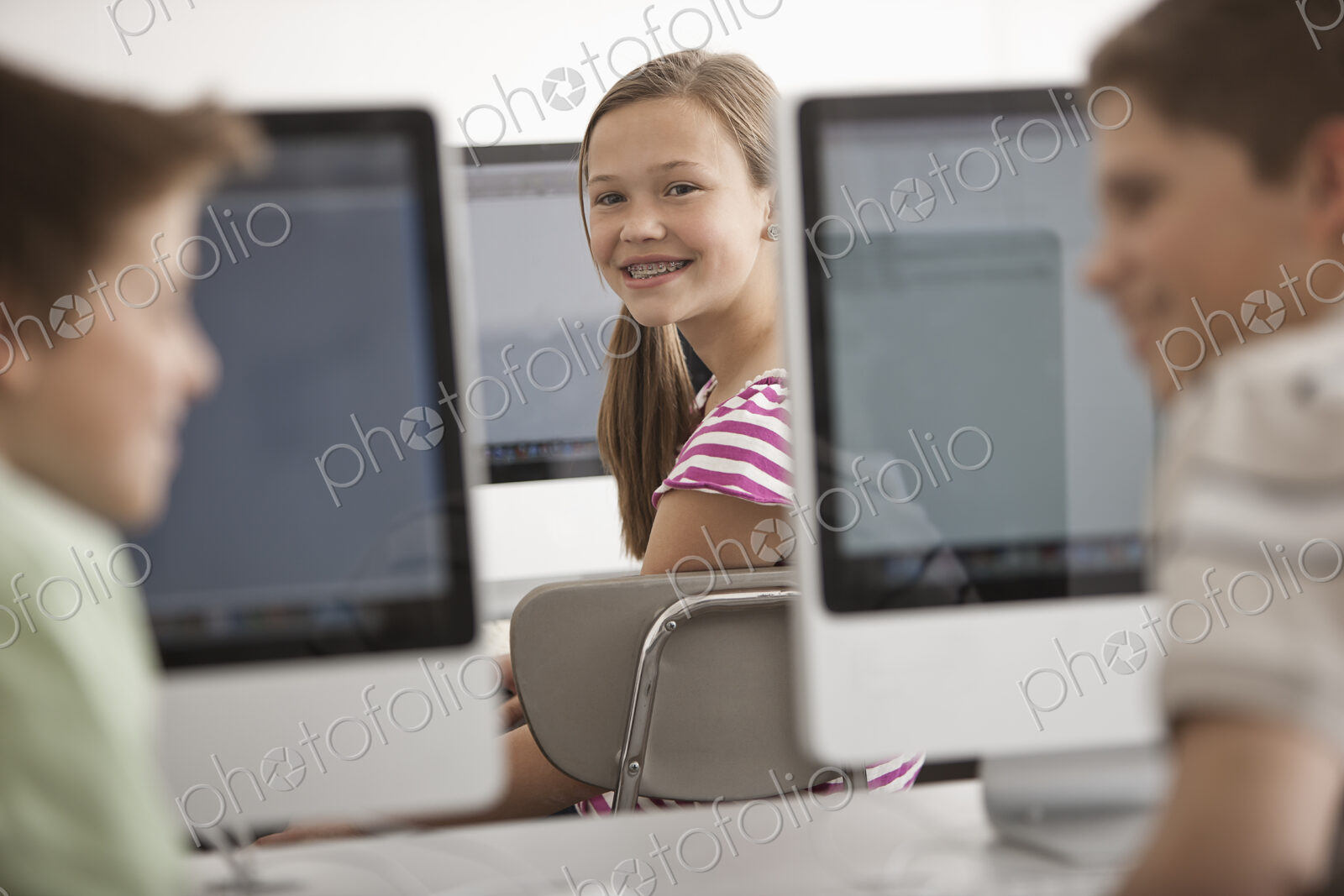 A school room computer lab with rows of computer monitors and seating. Three young…