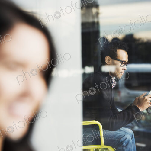 A seated man seen through a window looking at a cellphone, with an out-of-focus woman… A seated man seen through a window looking at a cellphone, with an out-of-focus woman…