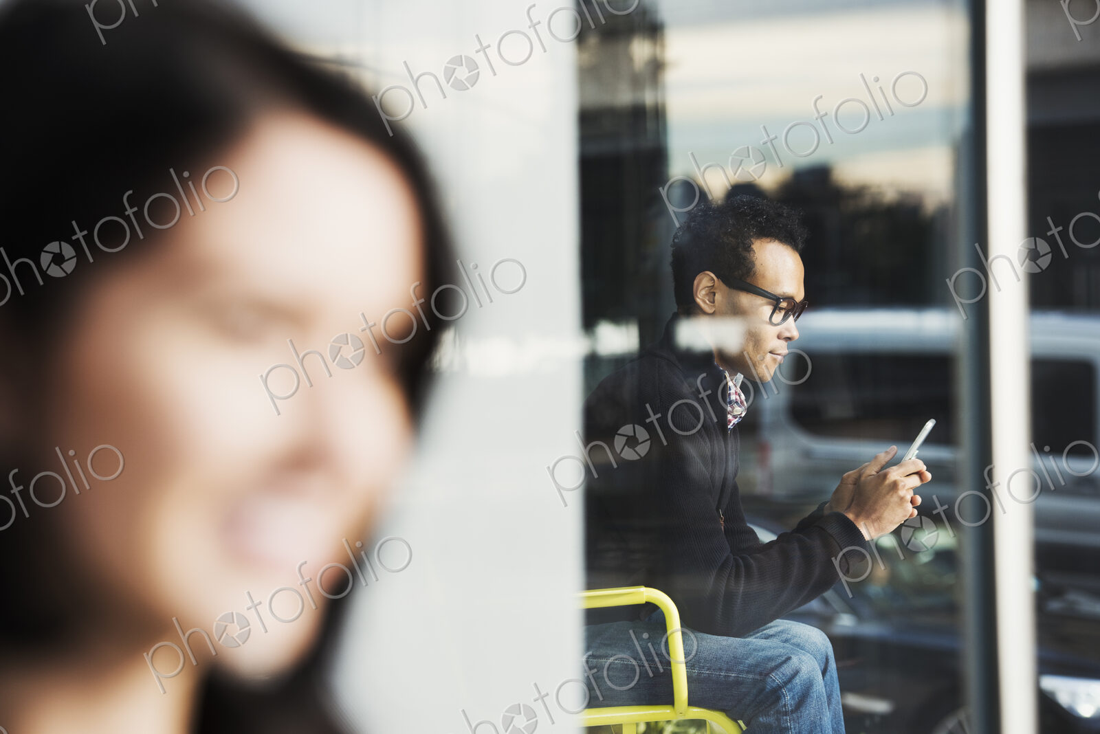 A seated man seen through a window looking at a cellphone, with an out-of-focus woman…