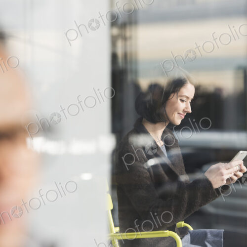 A seated woman seen through a window looking at a cellphone, with an out-of-focus man… A seated woman seen through a window looking at a cellphone, with an out-of-focus man…
