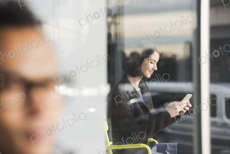 A seated woman seen through a window looking at a cellphone, with an out-of-focus man…