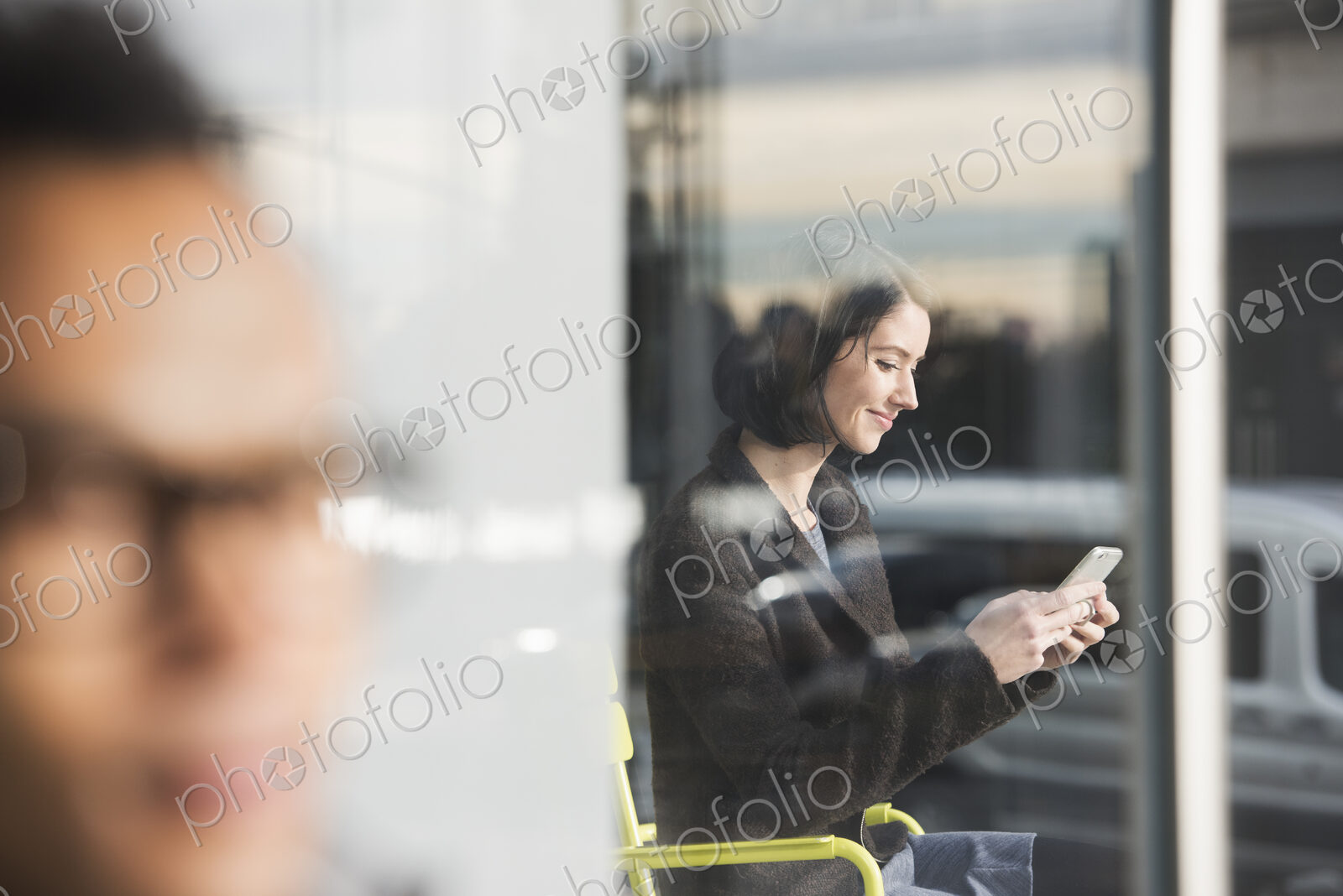 A seated woman seen through a window looking at a cellphone, with an out-of-focus man…