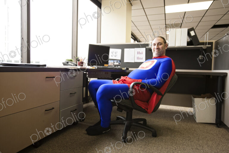 A self satisfied caucasian businessman super hero sits in his office cubicle.