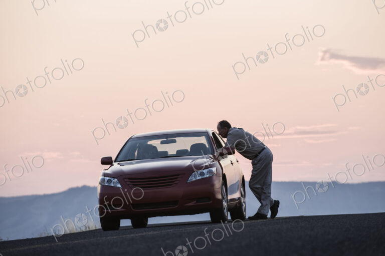 A senior Caucasian man leaning in a car window chatting with a person in the…