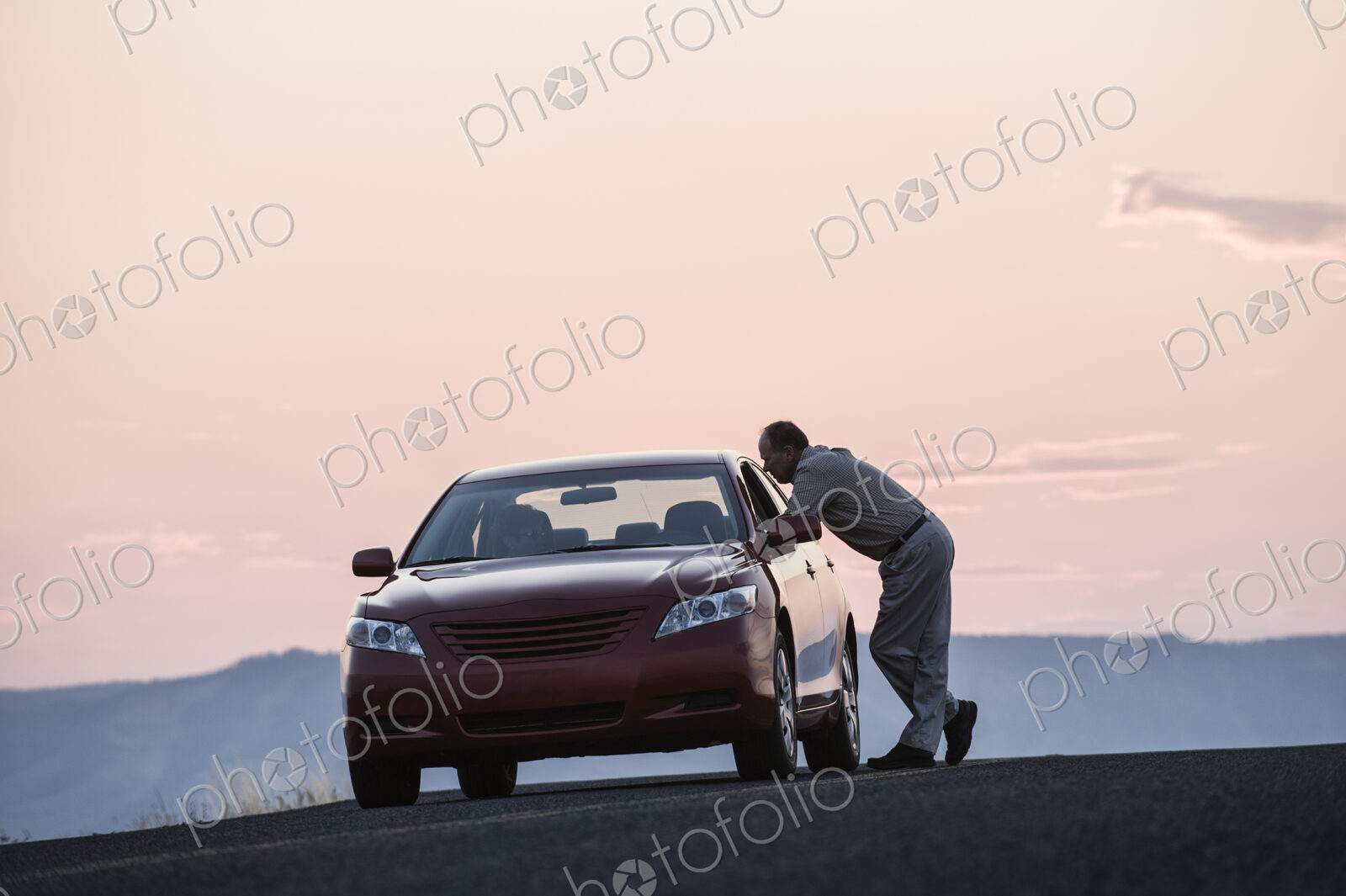 A senior Caucasian man leaning in a car window chatting with a person in the…