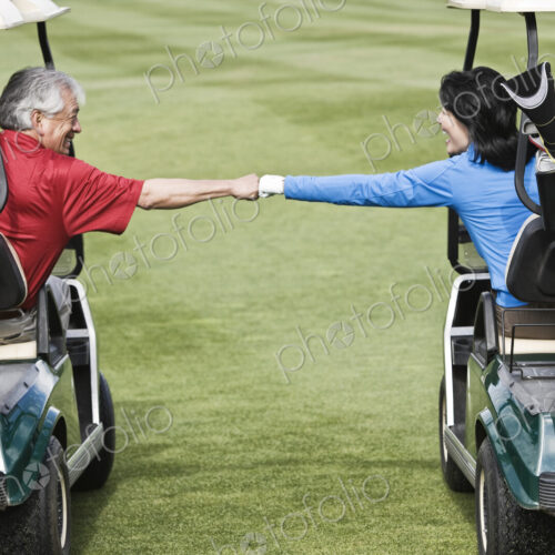 A senior couple doing a fist bump between golf carts on the fairway of a golf course. A senior couple doing a fist bump between golf carts on the fairway of a golf course.