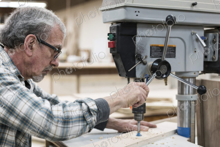 A senior man with glasses and beard in a woodworkers shop, using a machine.