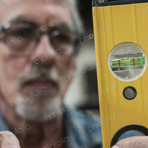 A senior man with glasses and beard in a woodworkers shop, using a spirit level…