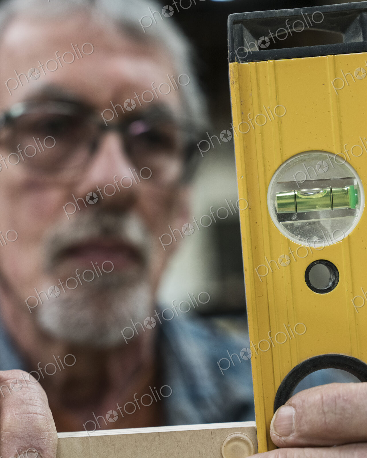 A senior man with glasses and beard in a woodworkers shop, using a spirit level…