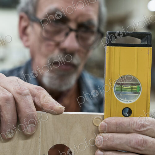 A senior man with glasses and beard in a woodworkers shop, using a spirit level…