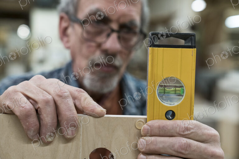A senior man with glasses and beard in a woodworkers shop, using a spirit level…