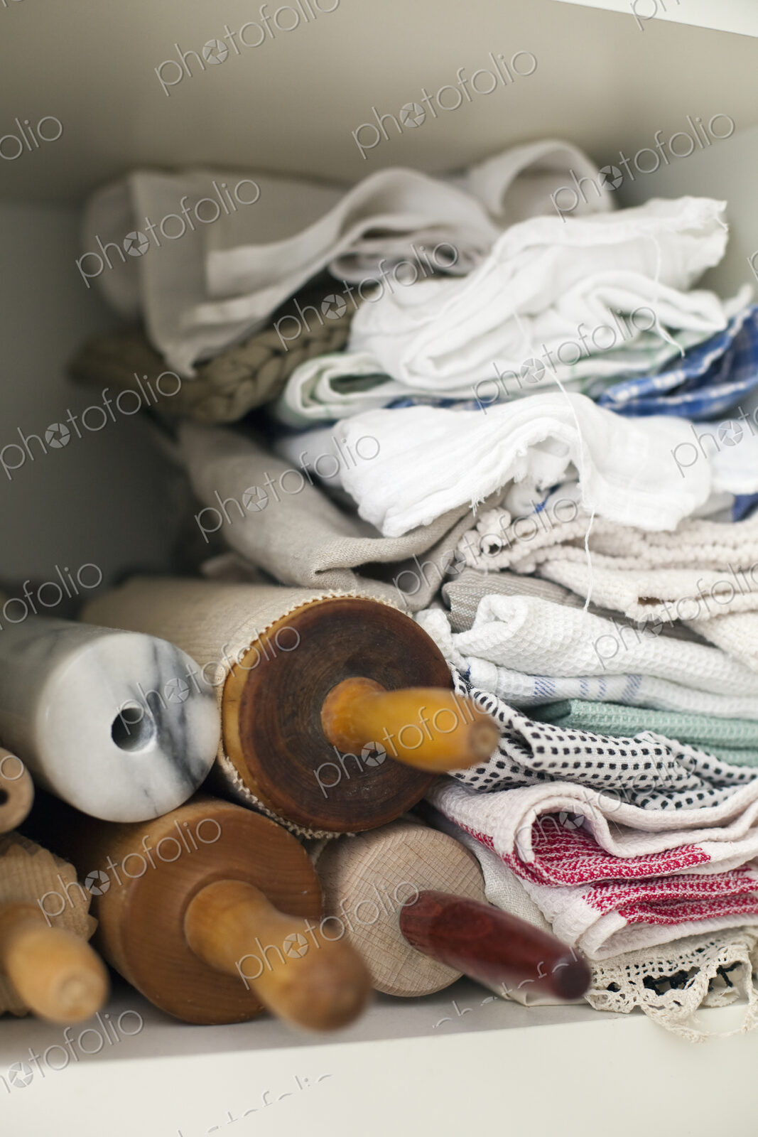 A shelf piled high with cloths, linen and a small stack of rolling pins or wooden…