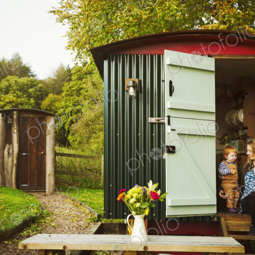 A shepherd's hut with open door beside a path to a small rustic shed, and a woman with…