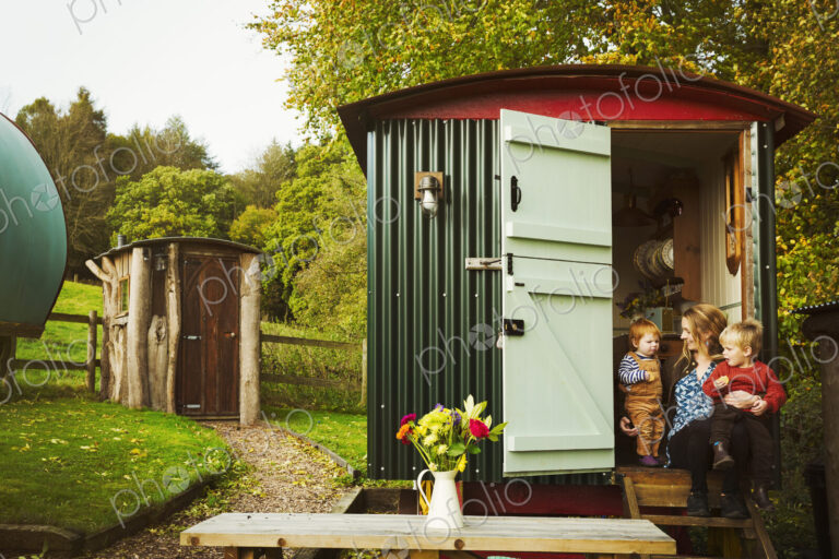 A shepherd's hut with open door beside a path to a small rustic shed, and a woman with…