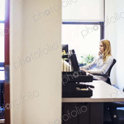 A shot of the doorway to two offices, a young woman sitting at a desk in one, and a… A shot of the doorway to two offices, a young woman sitting at a desk in one, and a…