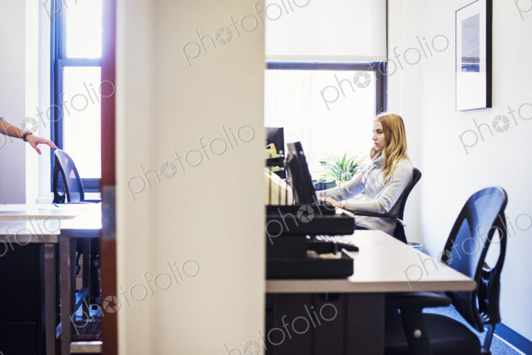 A shot of the doorway to two offices, a young woman sitting at a desk in one, and a…