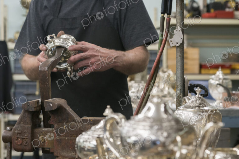 A silversmith in his workshop holding a silver pot examining it.