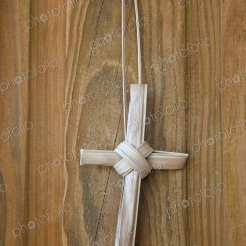 A simple handmade cross crafted from dried palm fronds hangs against a rustic wooden…