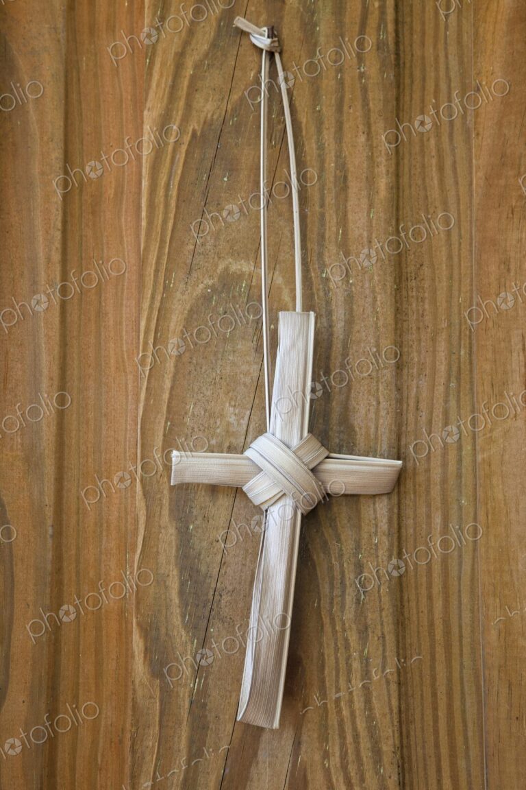 A simple handmade cross crafted from dried palm fronds hangs against a rustic wooden…