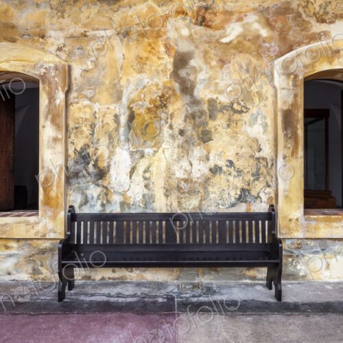 A simple wooden bench sits between two arched windows against a textured, weathered… A simple wooden bench sits between two arched windows against a textured, weathered…