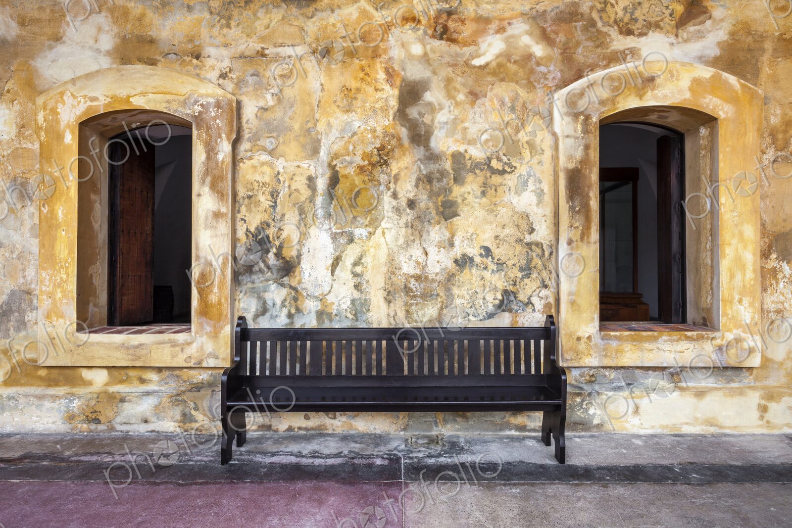 A simple wooden bench sits between two arched windows against a textured, weathered…