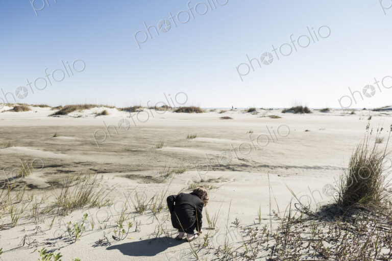 A six year old boy in soft white sand dunes, bending down to inspect something.