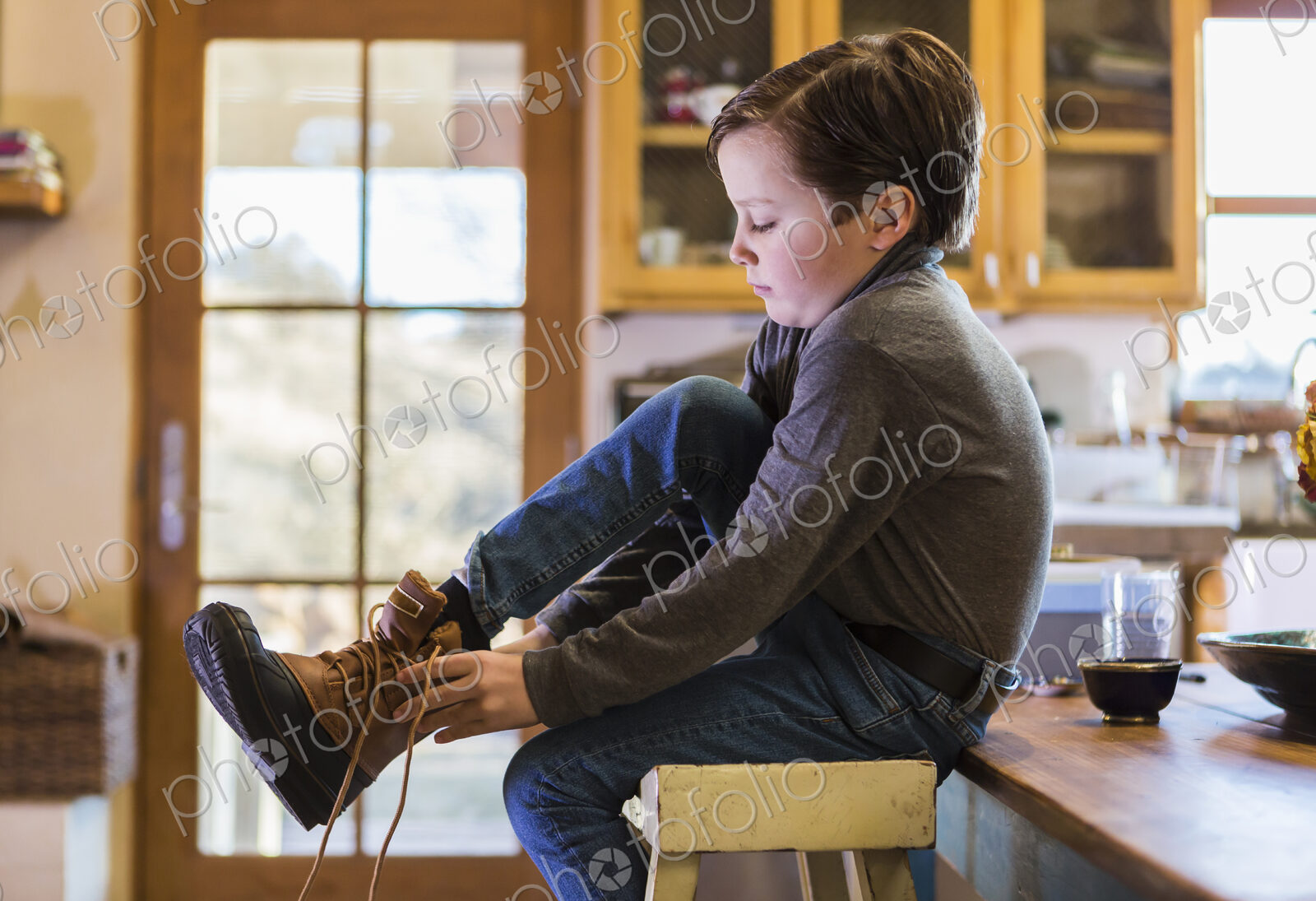 A six year old boy putting his boots on, sitting on a high stool.