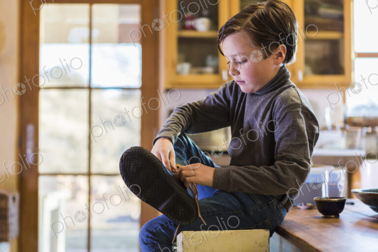 A six year old boy putting his boots on, sitting on a high stool.