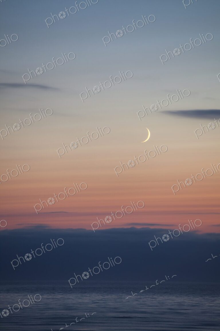 A slender crescent moon rises above the calm Pacific Ocean at dusk, Winchester Bay,…