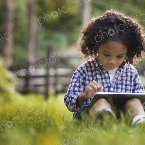 A small boy sitting on the grass using a tablet computer.